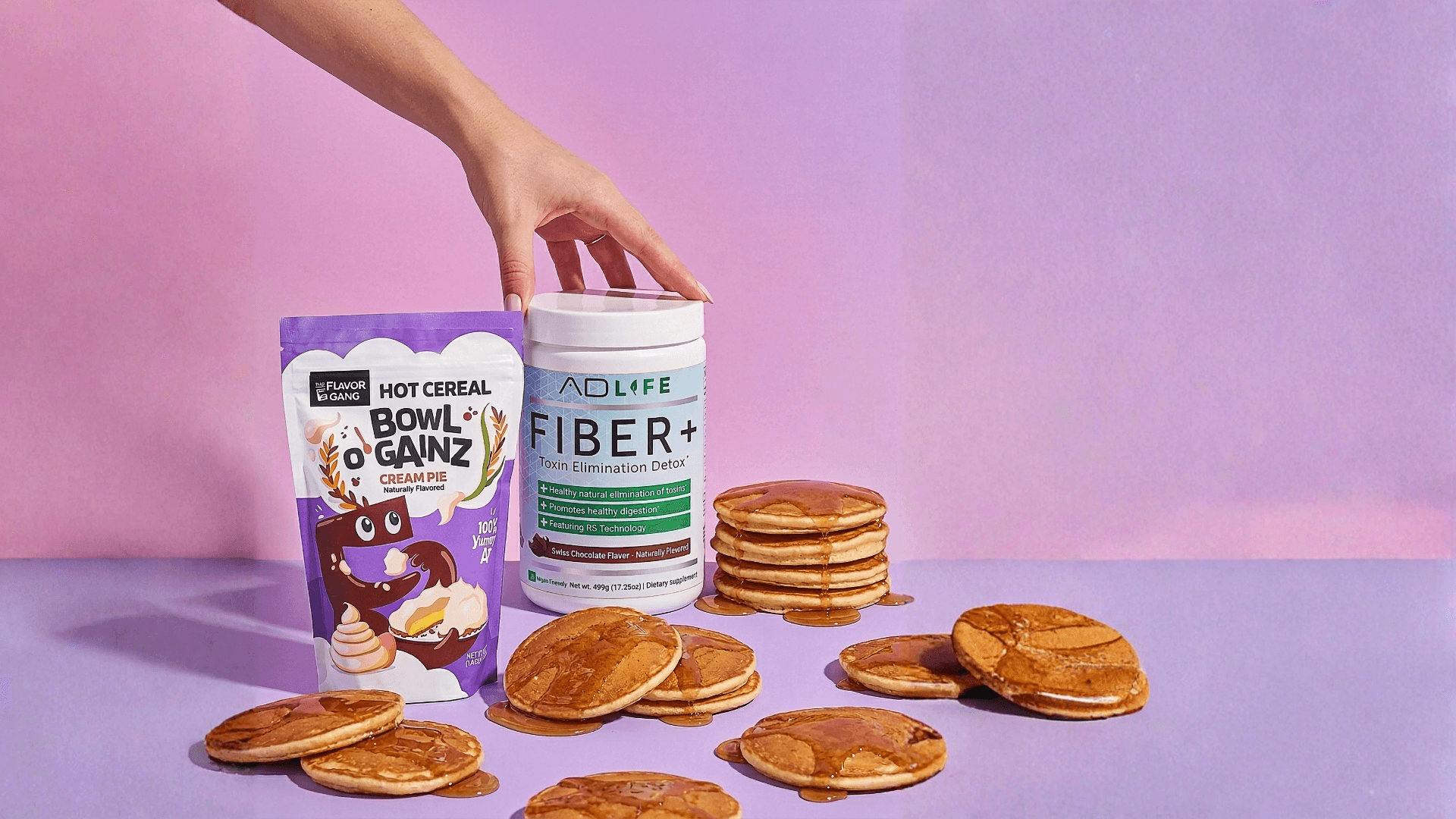 A hand places a white tub of AD LIFE Fiber+ beside a purple bag of The Flavor Gang Hot Cereal Bowl O’ Gainz Cream Pie on a pastel pink-purple background, surrounded by syrup-drizzled pancakes.